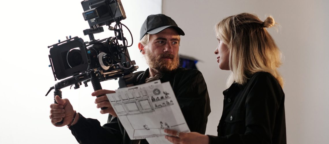 Young bearded cameraman and his assistant with documents consulting about shooting while standing in front of camera in studio